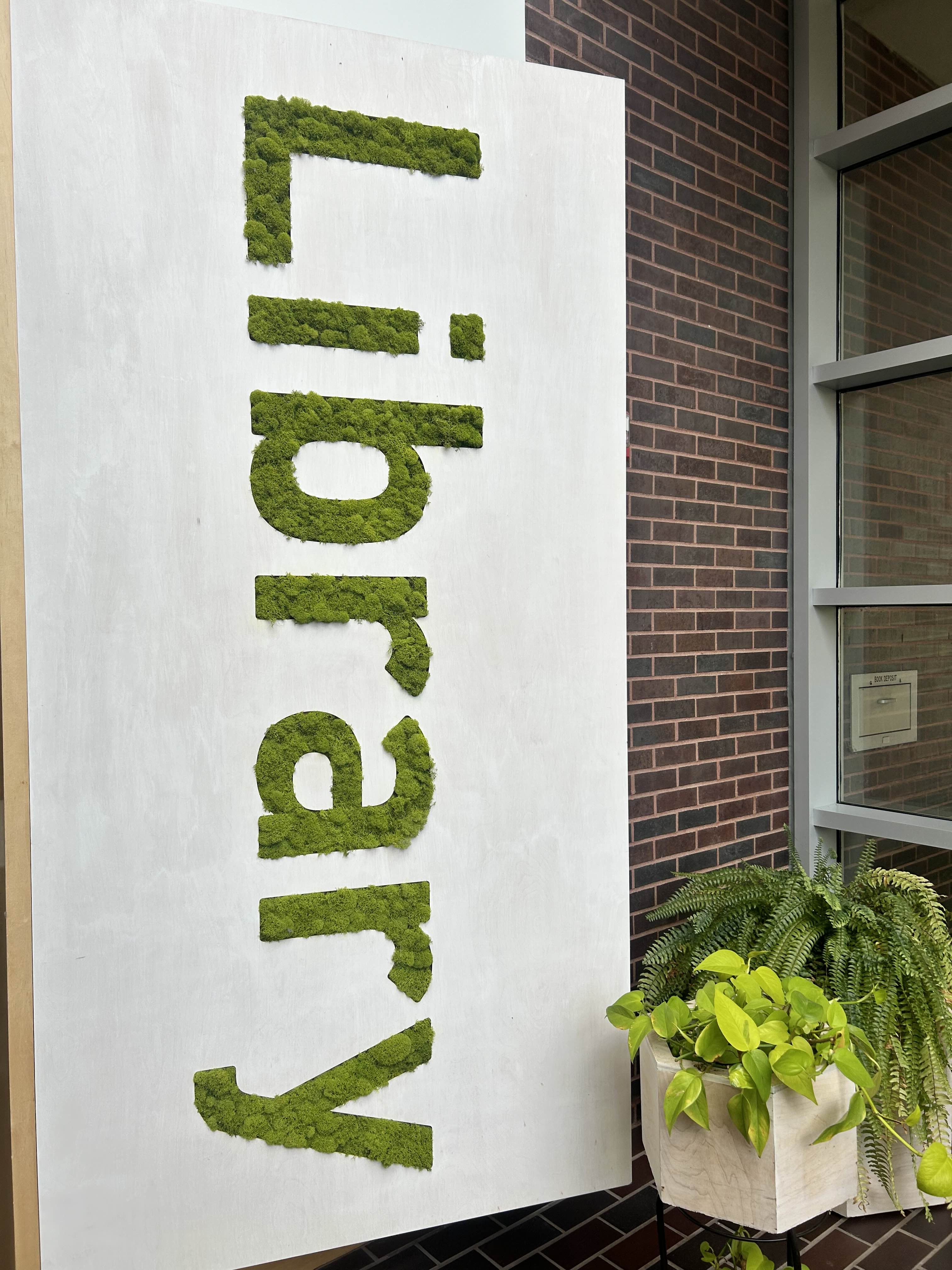 Picture of a sign made with the letters made of fake green moss that says 'Library' next to two potted plants. 